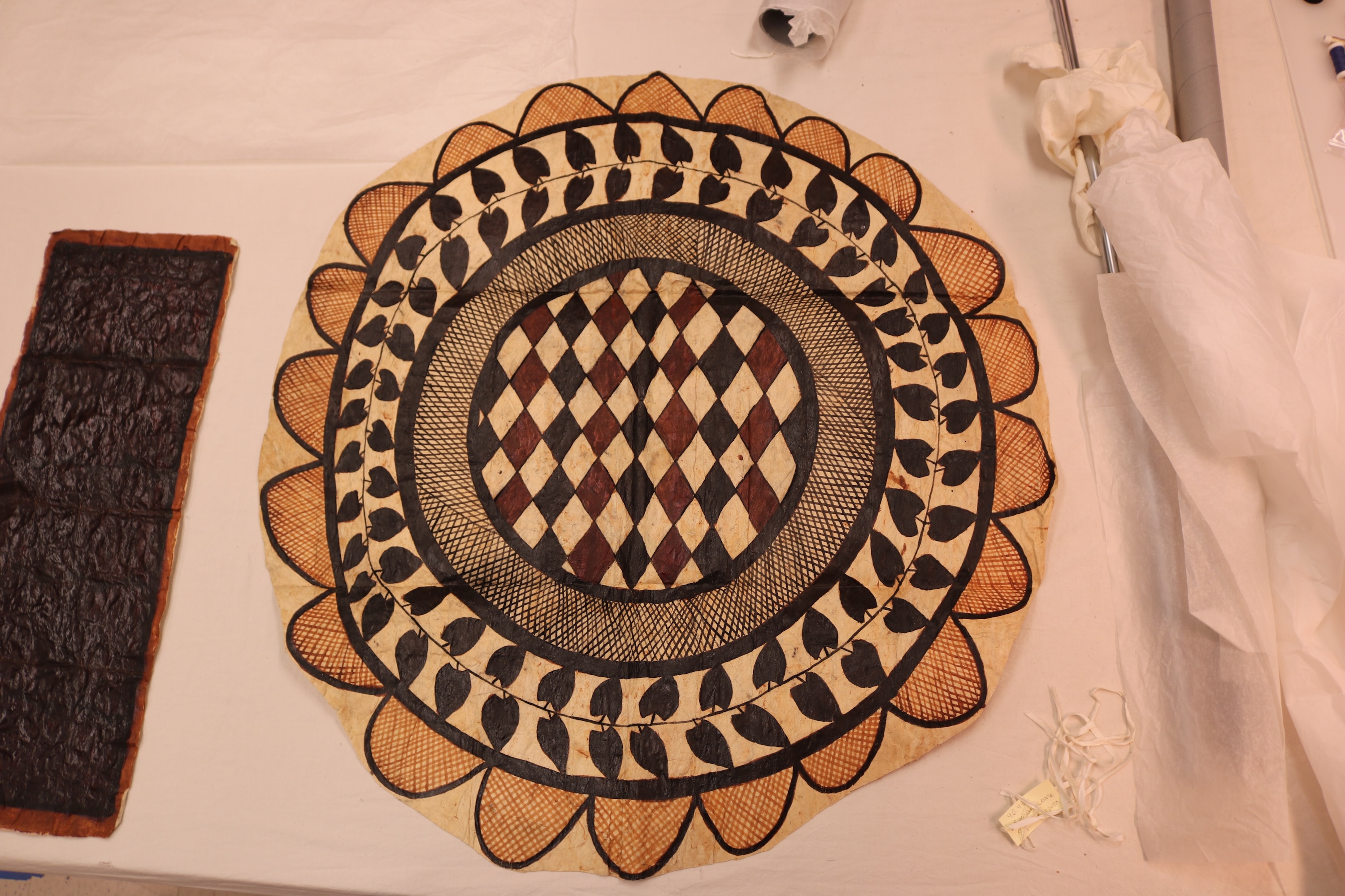 A circular siapo with a central checkerboard pattern, a border of leaves, and a scalloped design around the edge